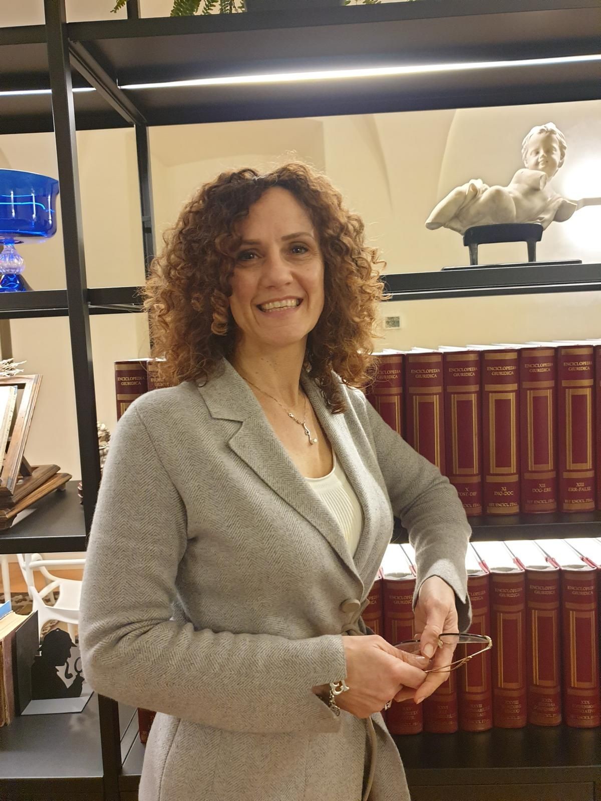 Woman smiling, standing near bookshelves with red books and decorative items, holding glasses in hands.